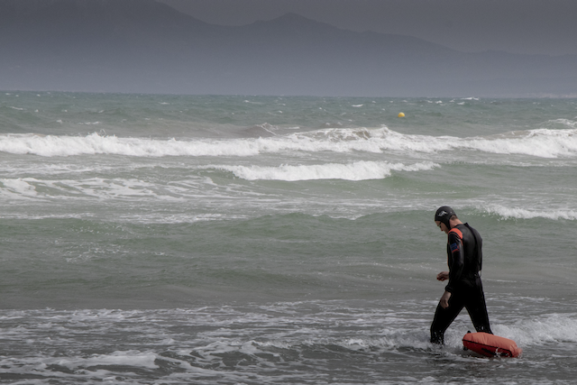 Ein Surfer mit rotem Board geht in die Brandung, im Hintergrund liegen Berge.